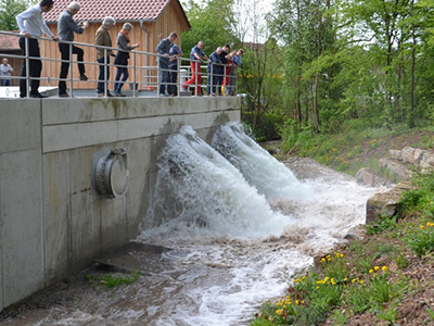 Auf dem Hochwasserentlastungspumpwerk Oppenweiler beobachten Personen den Probelauf des Beckens. Aus zwei von drei Auslässen läuft das Wasser in das Gewässer. 
