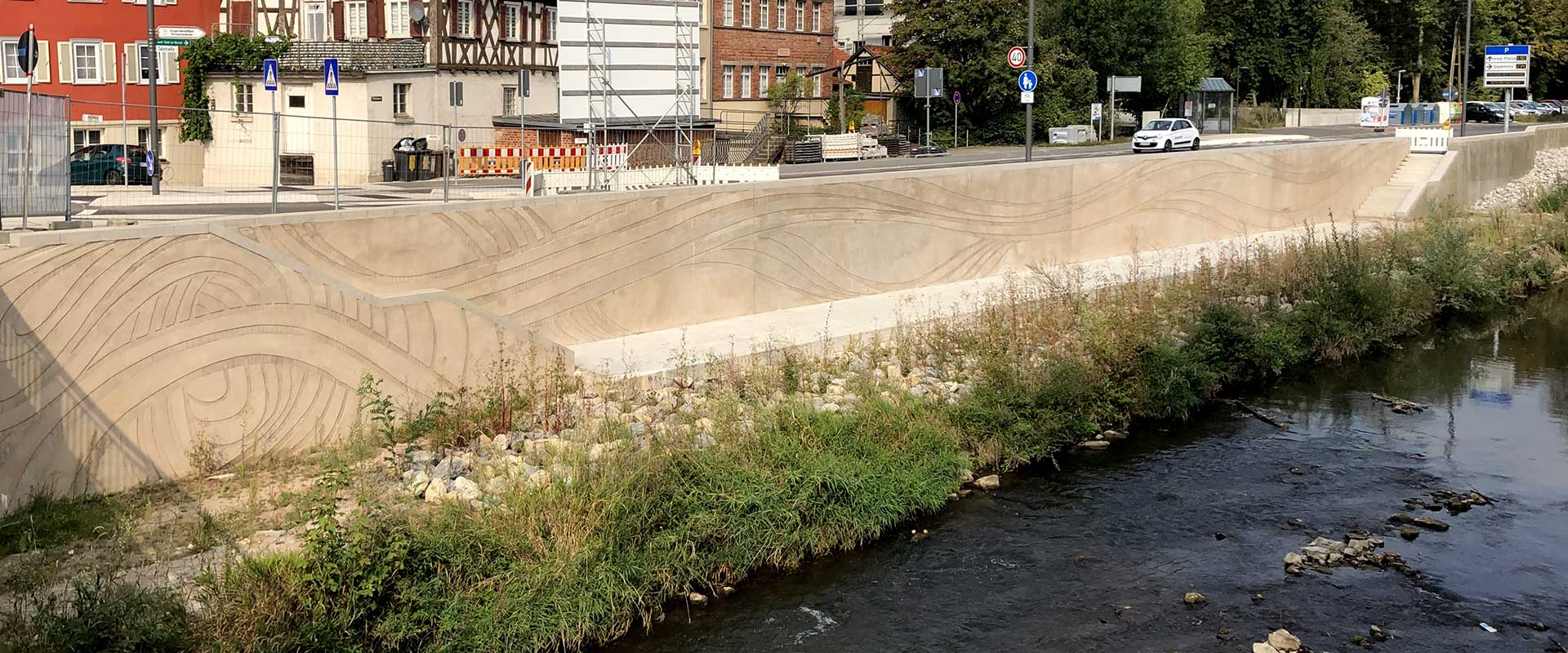 Es ist die neue wellenartig gestaltete Hochwasserschutzmauer im Bereich des neuen Tiefufers an der Murr in der Backnanger Talstraße nach der Aspacher Brücke zu sehen. Im Hintergrund sieht man noch die Baustellensicherung und das Baustellen-Hinweisschild zu dieser Maßnahme.