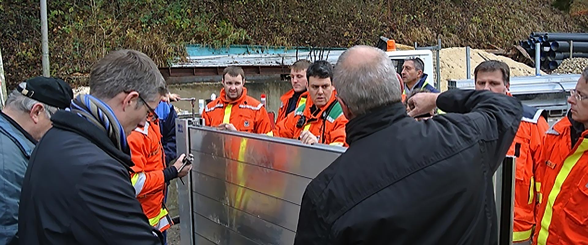 Mehrere Einsatzkräfte hören den Ausführungen zum mobilen Hochwasserschutzsystem in Oppenweiler zu. Ein Mann zeigt bei der Einführung ein mobiles Hochwasserschutzmauerelement.